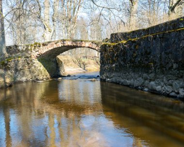 Kuja nehri üzerindeki Masonry köprüsü, açık mavi gökyüzü ile güneşli bir bahar günü, geçen yılın çimleriyle kaplanmış küçük bir nehrin kıyıları. Sudaki ağaç yansımaları, Kuja nehri, Letonya