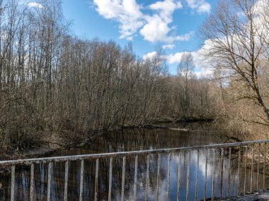 Köprüden nehir manzarası, ilkbaharın başlarında küçük bir nehir, mavi gökyüzü ve sudaki yansımalar, yapraksız ağaçlar, güneşli bahar manzarası, Ruja nehri, Letonya