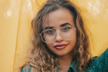 Arka planda sarı bez üzerine gözlük takan genç hippi modern kadın. Kıvırcık kahverengi saçlı.