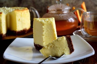 Beyaz tabakta bir dilim cheesecake ve gri arka planda bir fincan çay.
