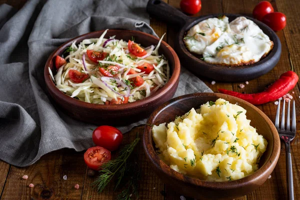 Hearty lunch: mashed potatoes in a wooden bowl, vitamin salad of vegetables in a clay plate, fried eggs on a wooden platter