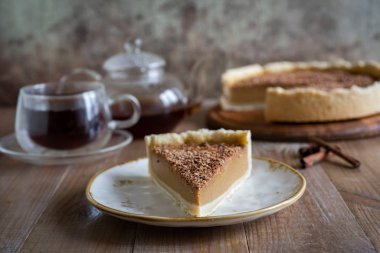 Ahşap bir masada eski bir tabakta lezzetli bir dilim kabaklı cheesecake. Arka planda balkabaklı peynirli kek ve bir bardak sıcak çay var.