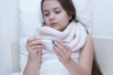 Kız termometreye bakıyor. Çocuğun koltukaltında termometre var. Yatakta küçük bir kız, sıcaklığı ölçüyor, üzgün bir yüz, kızın boynunda bir atkı ve koltuk altında bir termometre var.