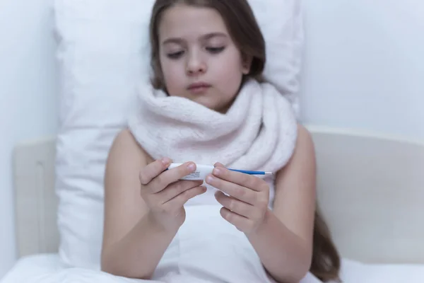 girl looks at a thermometer. the child is sick with a thermometer under the armpit. A little girl in the bed, measures the temperature, a sad face, the girl has a scarf around her neck and a thermometer under her armpit
