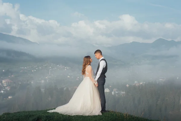 Pareja joven de bodas despertando en la colina en las montañas con hermoso paisaje y niebla en el fondo al amanecer. Vista posterior elegante pareja de novia y novio en un paseo de bodas —  Fotos de Stock