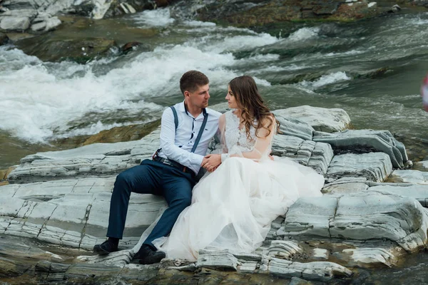 Hermosa pareja de boda besándose y abrazándose cerca de la orilla de un río de montaña con piedras —  Fotos de Stock