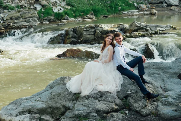 Schönes Hochzeitspaar küsst und umarmt sich am Ufer eines Gebirgsflusses mit Steinen — Stockfoto