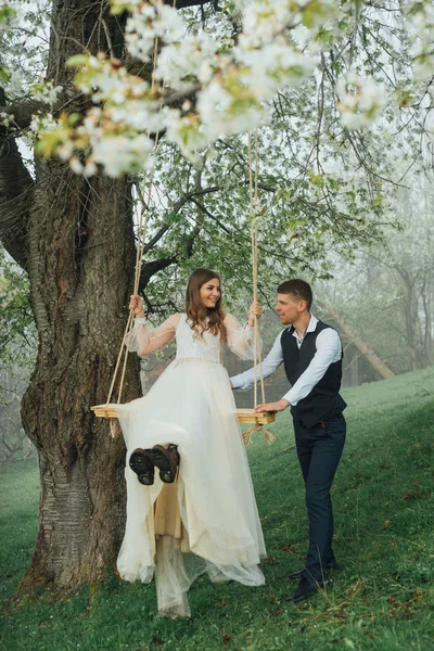 Braut und Bräutigam reiten auf einer Seilschaukel im Garten. Hochzeit im Wald. — Stockfoto