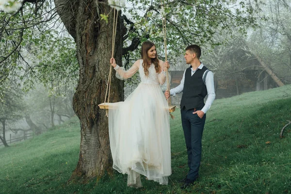 Braut und Bräutigam reiten auf einer Seilschaukel im Garten. Hochzeit im Wald. — Stockfoto