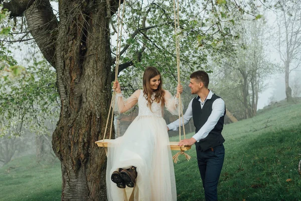 Ein stilvoller Bräutigam rollt die Braut auf einer Schaukel vor dem Hintergrund der wunderschönen Natur und des grünen Rasens. fröhliche Zeit bei der Hochzeit. Lustige Frischvermählte albern herum. — Stockfoto