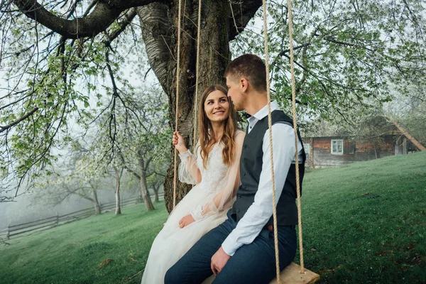 Braut mit der perfekten Figur sitzt auf der Schaukel, der Ehemann daneben. Außenaufnahmen, Frühlingspark oder Sommerkonzept. Liebe, sinnliche Menschen — Stockfoto