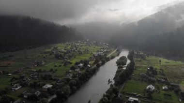 İçerideki sislerin içinden uçan, yukarıdan aşağıya doğru uçan hava aracının görüntüsü, cennetlik köyü, sırada kulübeleri, arka planda yeşil dağları ve nehri gözler önüne seriyor..