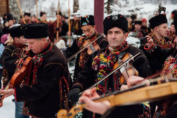 Kryvorivnia, Ukrajna - 2019. január 7.: Nemzeti hutul jelmezes hegedűművész hagyományos régi dalokat játszik, míg más férfiak az ukrán Kárpátok karácsonyi ünnepségén énekelnek. — Stock Fotó