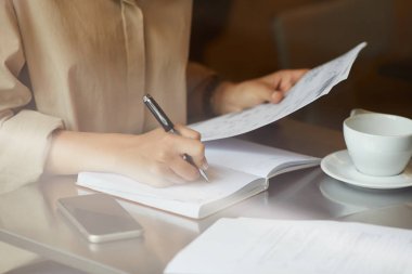 Genç iş kadınının elinde belge tutarken ve bir fincan kahveyle masada otururken not defterine gelecekle ilgili planlar yazarken.