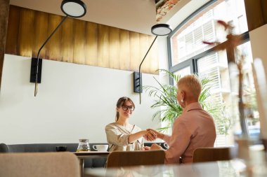 Genç iş kadını, masada oturmuş, kafedeki iş görüşmelerinde çalışma saatlerini tartışırken olgun iş kadınlarına el sıkışıyor.
