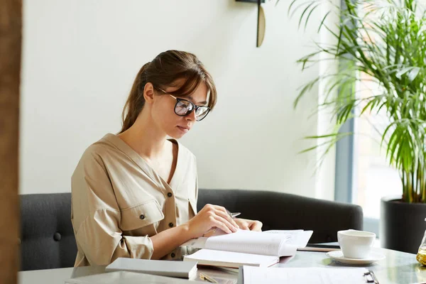 Allvarlig Ung Affärskvinna Glasögon Sitter Arbetsplatsen Och Tittar Anteckningar Anteckningsblock — Stockfoto