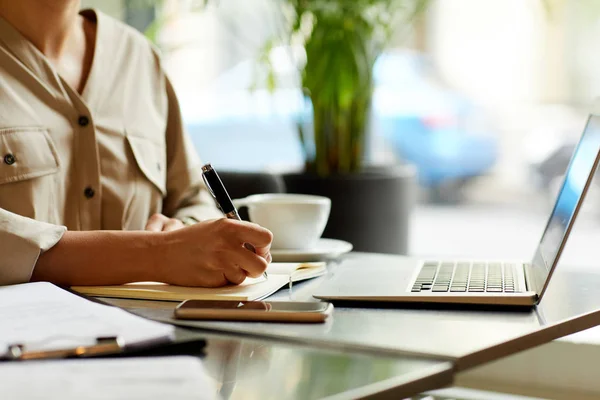 Laptopun önünde oturan ve kahve içerken not defterine notlar alan bir kadının yakın çekimi.