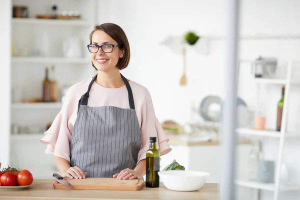 穿着眼镜和围裙的年轻女子站在厨房里切蔬菜时对着相机笑的画像 — 图库照片