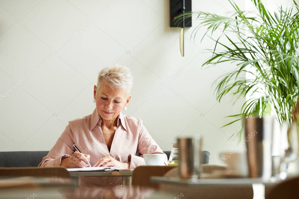 Serious mature businesswoman concentrating on work making notes at the table at office