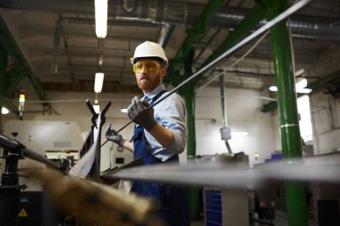 Manual worker in protective workwear and glasses wearing gloves holding iron pipe while working in the factory