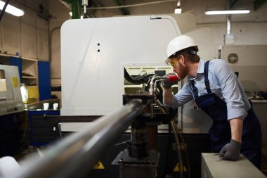 Young operator in protective glasses and workwear standing and working on lathe in the factory