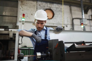 Serious bearded engineer in work helmet concentrating on work watching for the work of machine in the plant