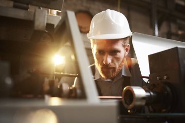 Serious bearded engineer in work helmet concentrating on work watching for the work of machine in the plant