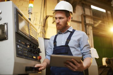Young operator in protective work wear using digital tablet while standing near the machine in the factory 