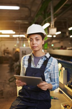 Female technician in work helmet controlling the work of machine with digital tablet in the plant 