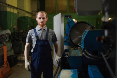 Portrait of young bearded mechanic in overalls looking at camera while standing in the factory 