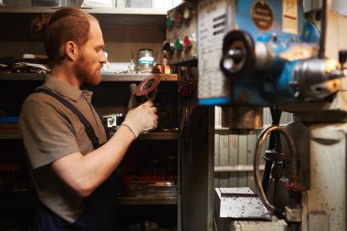 Young bearded mechanic examining metal details while working in warehouse 