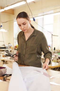 Genç ciddi terzi elinde makas tutuyor ve atölyedeki şablondan önce planı inceliyor.