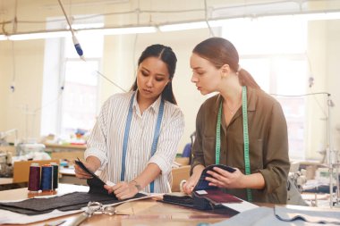 İki tasarımcı kumaş modeline bakıyor ve atölyedeki elbise koleksiyonunu seçiyor.