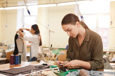 Ciddi bir terzi masada oturuyor ve arka planda mankenlerle çalışan bir meslektaşla kumaşları inceliyor.
