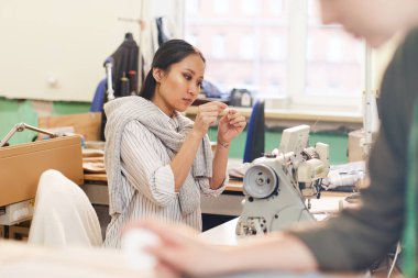 Ciddi bir Asyalı terzi atölyede dikiş makinesinin önünde otururken iğneyi ipliğe geçirir.