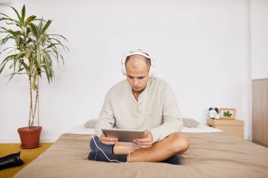 Kulaklıklı genç adam yatakta oturmuş yatak odasındaki dijital tabletten bir şeyler izliyor.