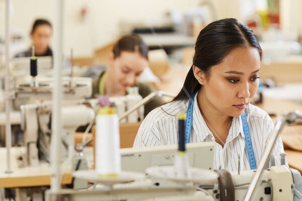 Серьезная азиатка, занимающаяся работой с швейной машинкой в мастерской с другими женщинами