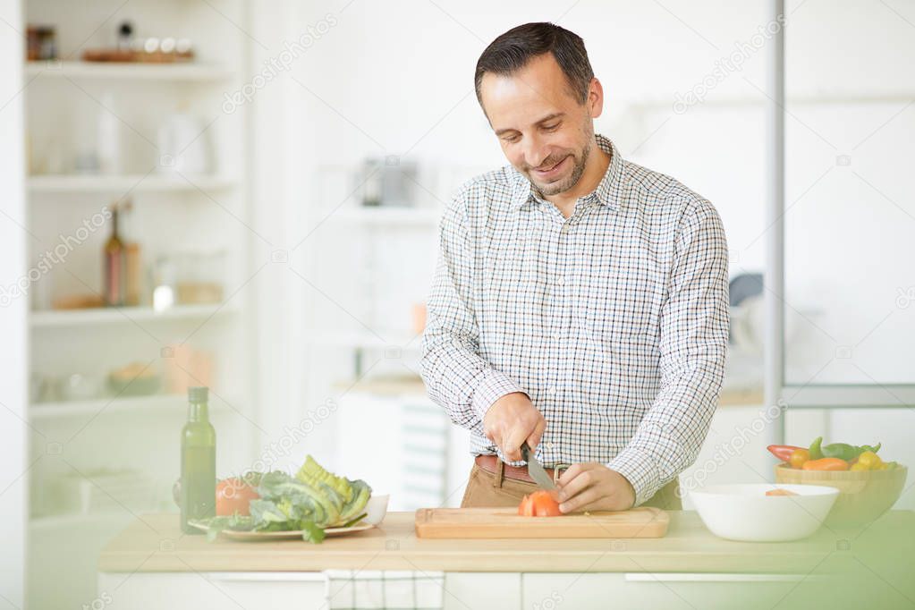 Young smiling man standing at the kitchen table and cutting vegetables on cutting board for salad in domestic kitchen