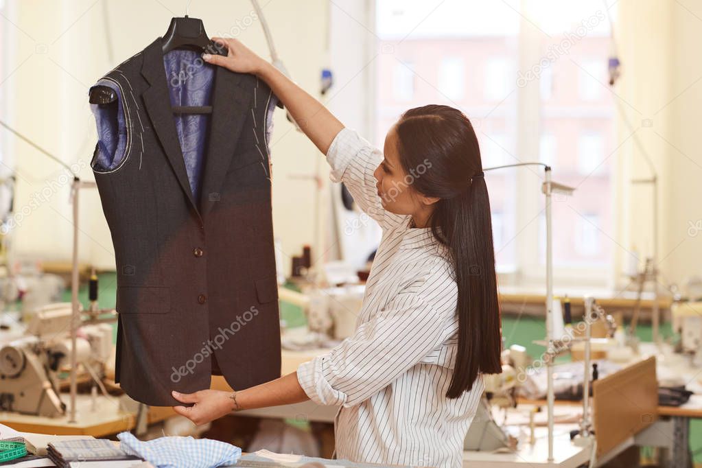 Asian young dressmaker holding unfinished suit in hands and examining it while working in workshop