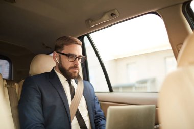 Young businessman in eyeglasses sitting on back seat of the car and working on laptop computer