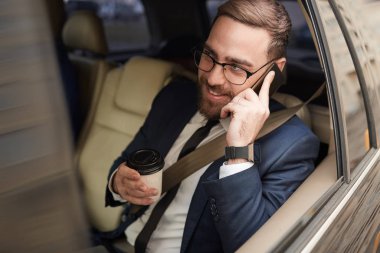 Young businessman in eyeglasses having conversation on the phone and drinking coffee while sitting on back seat in the car
