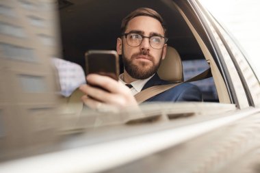 Serious young businessman using mobile phone and looking at view of the city while sitting in the car