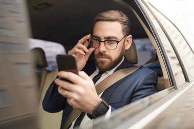 Young businessman in eyeglasses looking at screen of mobile phone while sitting on back seat of the car