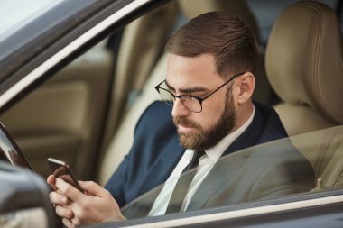 Young man in eyeglasses sitting in the car and texting message on mobile phone 
