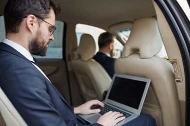 Young businessman in suit sitting on back seat of the car and working online using laptop while driver driving the car