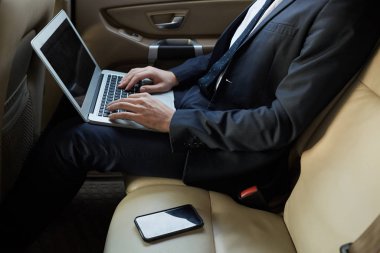 Close-up of businessman in suit sitting on back seat of luxury car and typing on laptop computer working online during driving