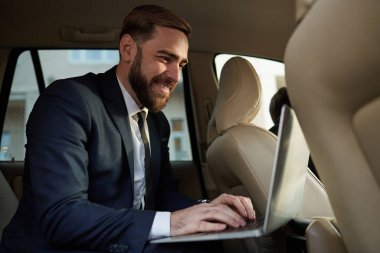 Young bearded businessman in suit sitting on back seat and typing on laptop computer  in taxi