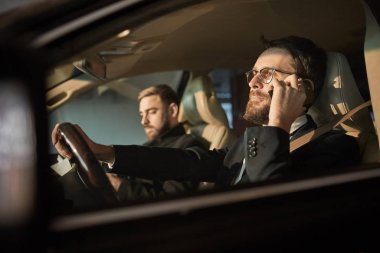 Young bearded driver in eyeglasses using his mobile phone during driving with passenger sitting near by him