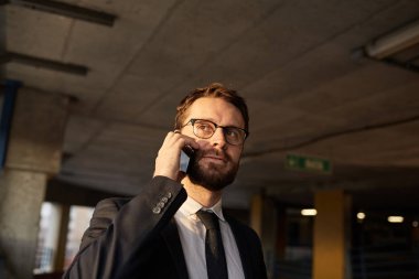 Young bearded businessman wearing eyeglasses and suit using mobile phone in the parking