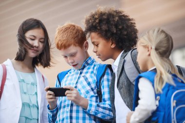 Red haired boy standing with mobile phone and laughing while talking to classmates outdoors